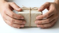 Hands protecting wrapped gift box with natural twine bow on white background. Eco-friendly packaging concept for holidays and special occasions.