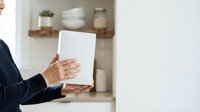 Mature woman in business attire holding blank tablet with open hand gesture in modern kitchen interior with shelving and decor.