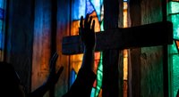 Silhouette of person reaching toward wooden cross with colorful stained glass window light creating dramatic spiritual atmosphere in church interior.