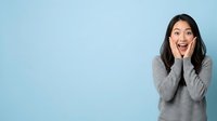 Excited young woman with dark hair and olive complexion wearing gray sweater, hands on cheeks, expressing surprise and joy against light blue background for advertising and lifestyle content.