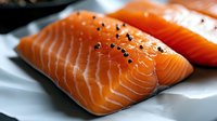 Fresh raw salmon fillets with black pepper seasoning on white paper, close-up view showing vibrant orange color and marbled texture for food photography.