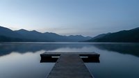Wooden dock extending into calm lake with mountain silhouettes at dawn, peaceful morning reflection creating serene landscape for meditation and relaxation.