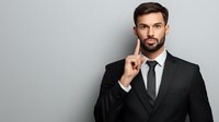 Young Caucasian businessman in black suit and tie with thoughtful expression holding finger near face against gray background, professional corporate portrait.