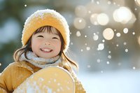 Young Asian girl in yellow winter coat and knit hat smiling brightly during snowfall, with soft bokeh lights and snowy background creating magical winter atmosphere.