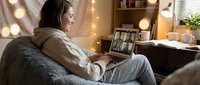 Woman in casual beige hoodie and jeans sitting on couch during video call on laptop in cozy home office with warm lighting and shelves.