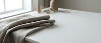 Minimalist bedroom interior with neatly folded beige linen blanket on white bed, natural sunlight streaming through window creating soft shadows on clean surfaces.