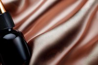 Black cosmetic bottle with dropper on luxurious brown silk fabric background with soft folds and waves creating elegant composition for beauty product advertising.