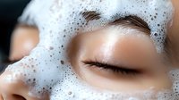 Close up view of young Asian woman with closed eyes and white foam cleanser on face during skincare routine and facial treatment at spa salon.