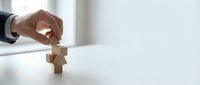 Male hand in business suit placing wooden blocks in cross or plus shape on white surface, symbolizing healthcare, medical services, or wellness strategy.