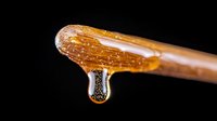 Golden honey dripping from wooden dipper against black background, macro photography showing crystalline texture and amber translucency in natural lighting.