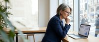 Mature businesswoman with glasses working on laptop in modern office, looking thoughtful while reviewing data by window with city view.