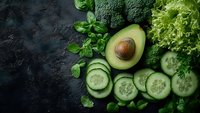 Fresh green vegetables including avocado, broccoli, cucumber slices and leafy greens arranged on dark slate background for healthy eating and nutrition.