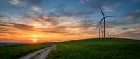 Wind turbines on green hill at sunset with dirt path leading through renewable energy farm generating clean electricity under dramatic orange sky.