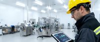 Male worker in yellow safety helmet and dark work jacket with reflective stripes holding tablet in modern industrial factory setting with machinery and equipment in background.