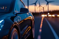 Modern car on highway at dusk with wind turbines silhouettes in background, dramatic sunset sky and bokeh lights reflecting on vehicle surface.