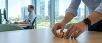 Professional businessman rolling wooden dice on office desk making strategic business decision with colleague working in background at modern workplace.