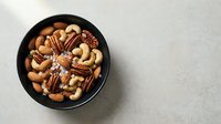 Mixed nuts with sea salt in a dark bowl on a light stone surface, top view with copy space for healthy snack and food concepts.