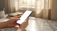 Person holding smartphone with blank white screen at marble desk near window with warm sunlight streaming through curtains for mockup design.