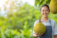 Young Asian female farmer holding fresh durian fruit in orchard garden, wearing apron and showing bright genuine smile against blurred green background.