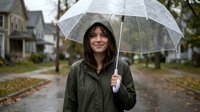Woman with dark wavy hair and olive complexion holding transparent umbrella on rainy residential street lined with autumn leaves and houses.