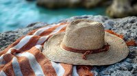 Straw sun hat with leather band resting on orange striped beach towel against rocky coastal background, capturing relaxed summer vacation mood.