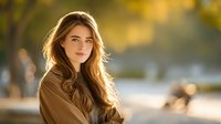 Young woman with long wavy hair illuminated by golden sunlight in autumn park setting, creating warm atmospheric portrait with soft bokeh background.