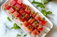 Fresh watermelon cubes on skewers garnished with bright mint leaves, served on white marble platter, summer fruit appetizer for party or picnic.