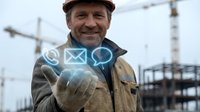Middle-aged male construction worker wearing hard hat and work jacket holding glowing digital communication icons at building site for business technology and contact concepts.