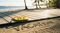 Yellow flip flops on a wooden boardwalk beside the word vacation written in sand, tropical beach with palm trees and ocean in background.