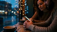 Young woman using smartphone in cozy cafe during evening with warm lighting and city bokeh background through window creating atmospheric mood.