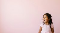 Young girl with dark hair and medium skin tone wearing a white shirt, looking upward with a thoughtful expression against a soft pink background, suitable for educational, parenting, and childhood development content.