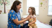 Female pediatric nurse in colorful scrubs examining young girl patient with teddy bear in cheerful medical office with nature wall decals.