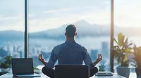 Asian businessman meditating in lotus position at desk with cityscape view through window, practicing mindfulness during work break in modern office space.