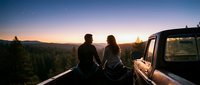 Couple sitting on vintage truck bed watching sunset over forested mountain valley at dusk, romantic travel and adventure lifestyle scene.
