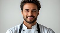 Confident male chef with beard and white uniform smiling against light background. Professional culinary portrait showing warmth and expertise.