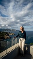 Mature blonde woman enjoying coastal view from modern terrace overlooking Mediterranean sea and mountains under dramatic cloudy sky.
