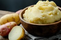 Creamy mashed potatoes served in rustic wooden bowl with raw potatoes on dark wooden table, comfort food close up on gray napkin background.