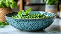 Fresh green peas garnished with mint leaves in textured turquoise ceramic bowl on marble surface, bright natural lighting creates appetizing food styling.