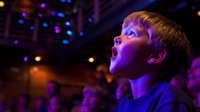 Young Caucasian boy with awestruck expression watching performance, illuminated by colorful stage lights in dark theater, face glowing with wonder and amazement.
