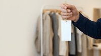 Close-up of hand holding blank white tag or label against blurred clothing rack background, suitable for retail, fashion, pricing, and product labeling concepts.