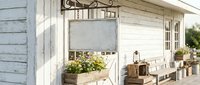 Cozy white wooden cottage porch with vintage furniture, potted plants and rustic decor elements creating charming farmhouse style outdoor living space.