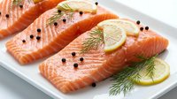 Fresh salmon fillets with lemon slices and dill garnish on white plate. Raw fish portions seasoned with black pepper ready for cooking or grilling preparation.