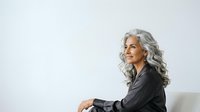 Mature woman with long gray hair wearing dark blazer, seated against white background, professional portrait for business and corporate use.