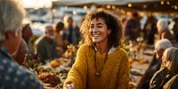 Young woman with curly hair laughing at outdoor dinner party, wearing yellow sweater among friends and family gathering at sunset.