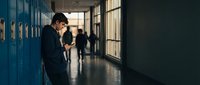 Sad teenage boy sitting alone in school hallway with blue lockers while other students walk by in background during daytime.