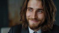 Young Caucasian man with long brown hair and beard smiling warmly at camera. Professional headshot with natural lighting showcases genuine expression and modern style.