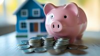 Pink ceramic piggy bank with stacks of silver coins on wooden surface, blue house model blurred in background, representing savings and home investment goals.