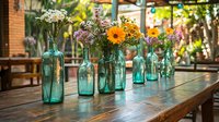 Colorful wildflowers in vintage turquoise glass bottles arranged on rustic wooden table, creating romantic garden party or wedding reception atmosphere in outdoor setting.