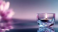 Lit candle in crystal glass holder creates serene ambiance with soft purple lighting and blurred floral background for relaxation and wellness concepts.