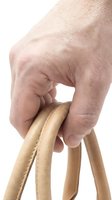 Caucasian hand gripping tan leather bag handles with visible stitching detail, close-up shot on white background for fashion and retail use.